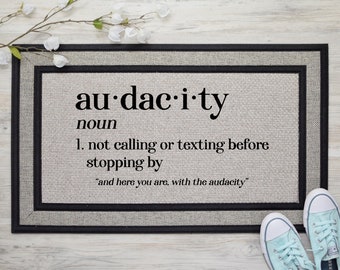May include: A doormat with the word "audacity" defined as "not calling or texting before stopping by". The doormat has a black border and a gray background.