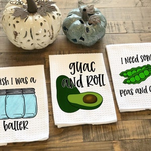 Op de afbeelding: Drie witte keukendoeken met grappige spreuken erop gedrukt. De eerste handdoek zegt "Ik wou dat ik een baller was" en heeft een afbeelding van drie glazen potten. De tweede handdoek zegt "guac and roll" en heeft een afbeelding van twee avocado's. De derde handdoek zegt "Ik heb wat erwten en rust nodig" en heeft een afbeelding van twee erwtenschoten.