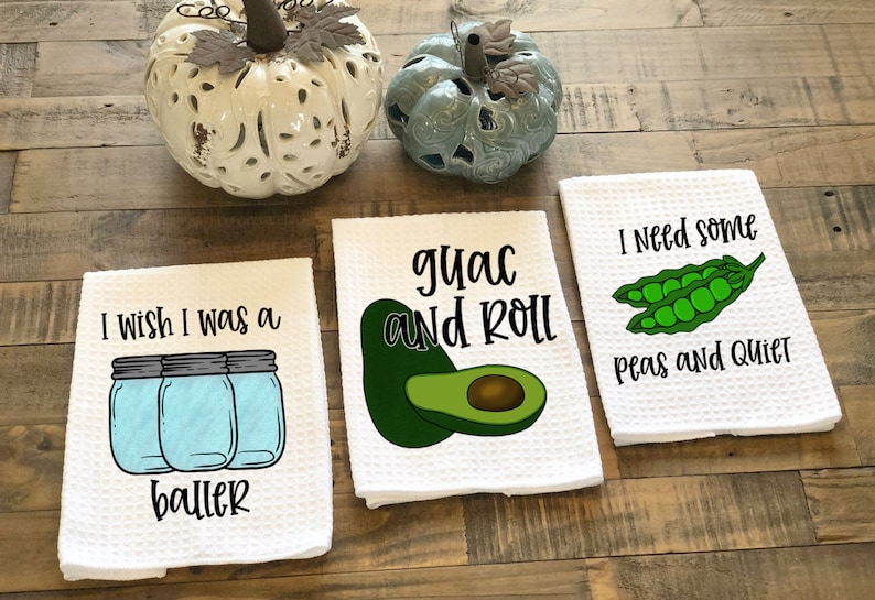 Op de afbeelding: Drie witte keukendoeken met grappige spreuken erop gedrukt. De eerste handdoek zegt "Ik wou dat ik een baller was" en heeft een afbeelding van drie glazen potten. De tweede handdoek zegt "guac and roll" en heeft een afbeelding van twee avocado's. De derde handdoek zegt "Ik heb wat erwten en rust nodig" en heeft een afbeelding van twee erwtenschoten.