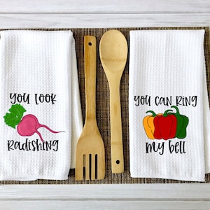 Op de afbeelding: Twee witte keukendoeken met grappige woordspelingen over eten. De eerste handdoek heeft een roze radijs met groene bladeren en de tekst "You look radishing". De tweede handdoek heeft drie paprika's, een gele, een rode en een groene, met de tekst "You can ring my bell".