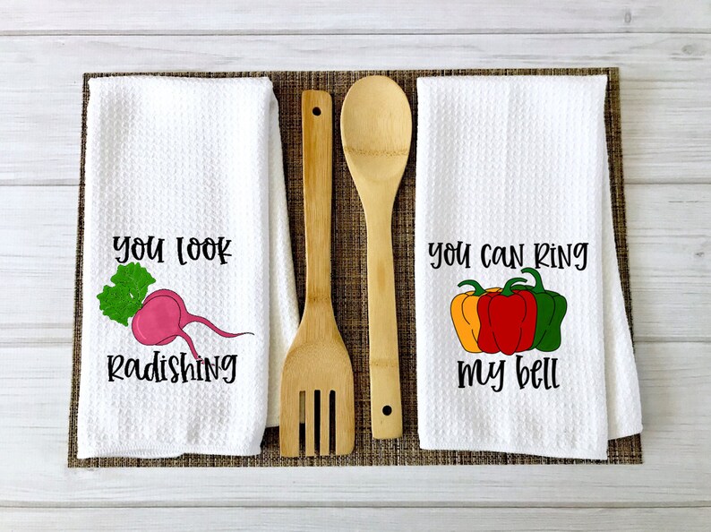 Op de afbeelding: Twee witte keukendoeken met grappige woordspelingen over eten. De eerste handdoek heeft een roze radijs met groene bladeren en de tekst "You look radishing". De tweede handdoek heeft drie paprika's, een gele, een rode en een groene, met de tekst "You can ring my bell".