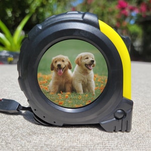 May include: A black and yellow tape measure with a photo of two golden retriever puppies on a green grassy field with yellow flowers. The puppies are looking at the camera and have their tongues sticking out.