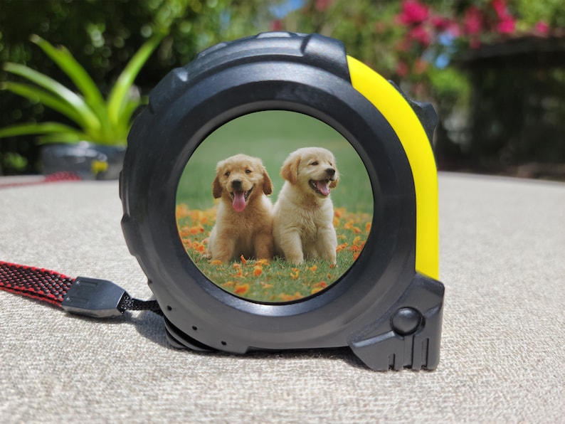 May include: A black and yellow tape measure with a photo of two golden retriever puppies on a green grassy field with yellow flowers. The puppies are looking at the camera and have their tongues sticking out.