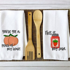 Op de afbeelding: Twee witte keukendoeken met spreuken erop. De eerste handdoek zegt "You've got a peach of my heart" met twee perziken erop. De tweede handdoek zegt "This is my jam" met een pot jam en een aardbei erop.