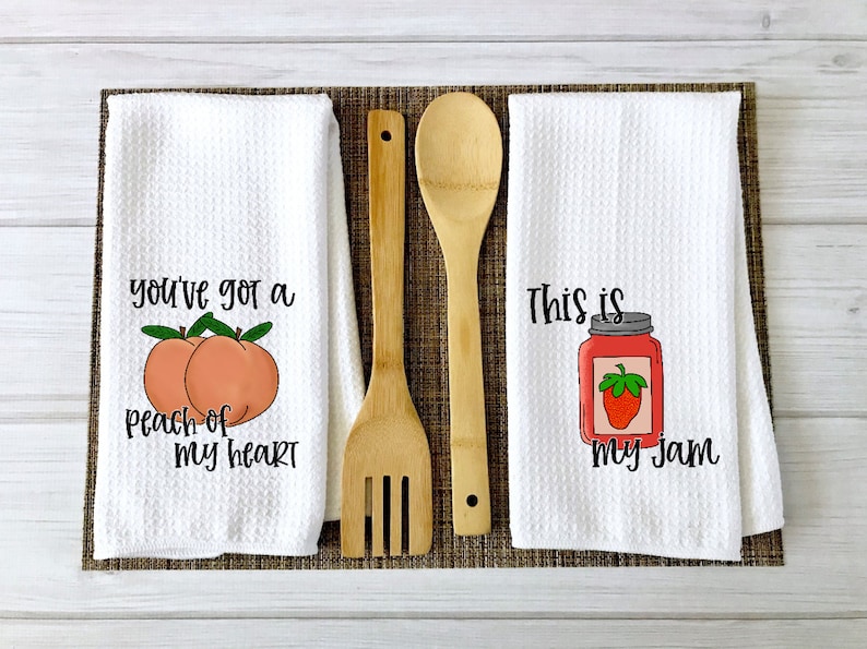 Op de afbeelding: Twee witte keukendoeken met spreuken erop. De eerste handdoek zegt "You've got a peach of my heart" met twee perziken erop. De tweede handdoek zegt "This is my jam" met een pot jam en een aardbei erop.