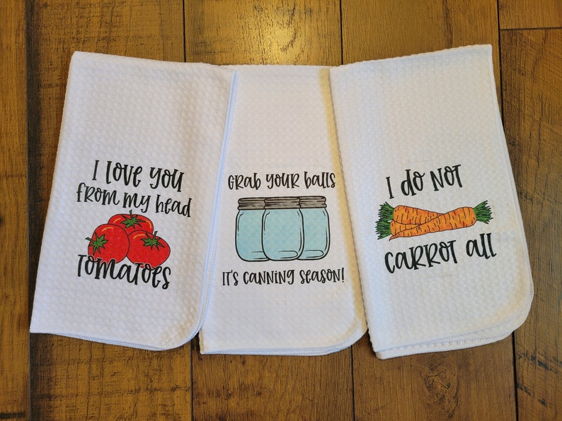 Op de afbeelding: Drie witte keukendoeken met humoristische spreuken erop gedrukt. De eerste handdoek zegt "Ik hou van je van mijn hoofd tot mijn tomaten". De tweede handdoek zegt "Pak je ballen, het is inmaaktijd!" met een afbeelding van drie glazen potten. De derde handdoek zegt "Ik ben niet helemaal wortel" met een afbeelding van drie wortels.