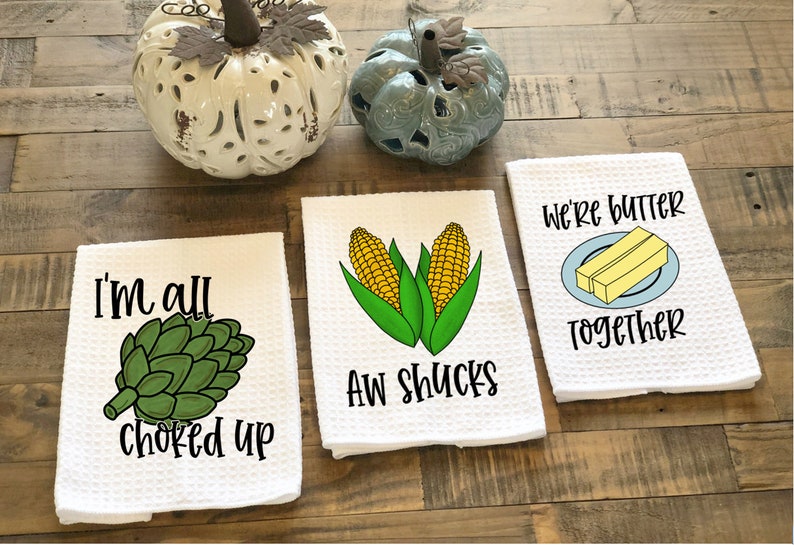 Op de afbeelding: Drie witte keukendoeken met verschillende woordspelingen met voedselthema. De eerste handdoek heeft een groene artisjok en de tekst "I'm all choked up". De tweede handdoek heeft twee ma&iuml;soren en de tekst "Aw shucks". De derde handdoek heeft twee stukjes boter op een blauw bord en de tekst "We're butter together".