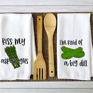 Op de afbeelding: Twee witte keukendoeken met grappige woordspelingen over eten. De eerste handdoek heeft een bos groene asperges en de tekst "Kiss my asparagus". De tweede handdoek heeft twee groene augurken en de tekst "I'm kind of a big dill".