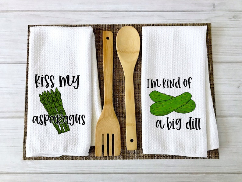 Op de afbeelding: Twee witte keukendoeken met grappige woordspelingen over eten. De eerste handdoek heeft een bos groene asperges en de tekst "Kiss my asparagus". De tweede handdoek heeft twee groene augurken en de tekst "I'm kind of a big dill".