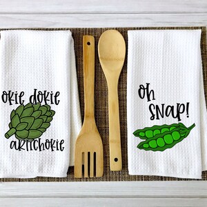 Op de afbeelding: Twee witte keukendoeken met een groene artisjok en de tekst "okie dokie artichokie" op de ene handdoek en groene erwten en de tekst "oh snap!" op de andere handdoek. Twee houten lepels bevinden zich in het midden van de afbeelding.