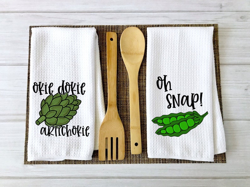 Op de afbeelding: Twee witte keukendoeken met een groene artisjok en de tekst "okie dokie artichokie" op de ene handdoek en groene erwten en de tekst "oh snap!" op de andere handdoek. Twee houten lepels bevinden zich in het midden van de afbeelding.