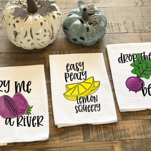 Op de afbeelding: Drie witte keukendoeken met kleurrijke illustraties van fruit en groenten en woordspelingen. De eerste handdoek zegt "Cry me a river" met een paarse ui. De tweede handdoek zegt "Easy peasy lemon squeezy" met een gele citroen. De derde handdoek zegt "Drop the beet" met een paarse biet.