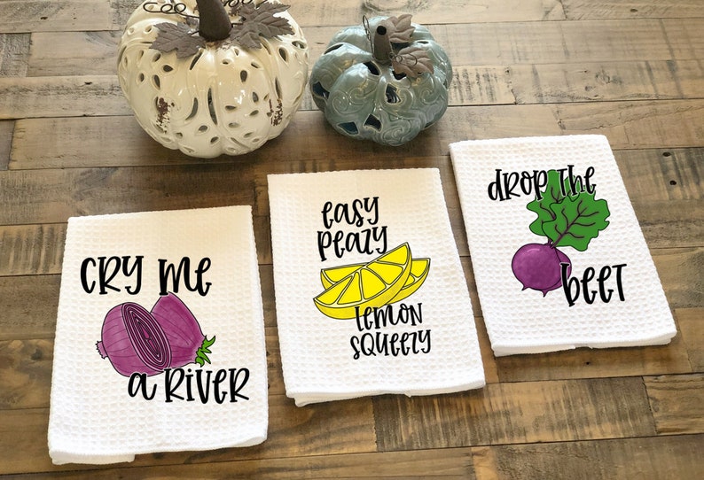 Op de afbeelding: Drie witte keukendoeken met kleurrijke illustraties van fruit en groenten en woordspelingen. De eerste handdoek zegt "Cry me a river" met een paarse ui. De tweede handdoek zegt "Easy peasy lemon squeezy" met een gele citroen. De derde handdoek zegt "Drop the beet" met een paarse biet.
