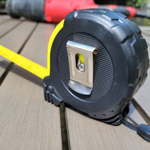 May include: A yellow and black tape measure with a metal hook and a black strap. The tape measure is on a wooden surface.