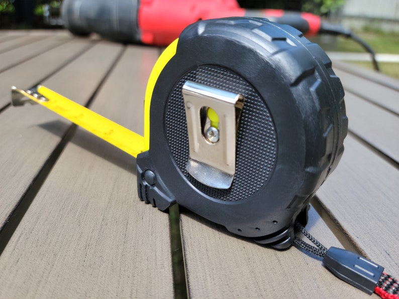 May include: A yellow and black tape measure with a metal hook and a black strap. The tape measure is on a wooden surface.