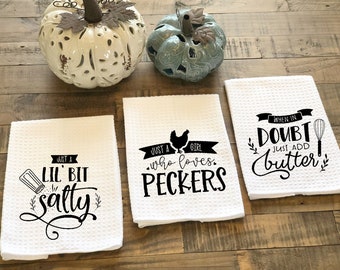 May include: Three white kitchen towels with black text. The first towel says "Just a Lil' Bit Salty" with a salt shaker graphic. The second towel says "Just a Girl Who Loves Peckers" with a chicken graphic. The third towel says "When in Doubt Just Add Butter" with a whisk graphic.
