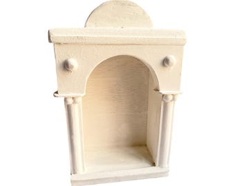 May include: A cream-colored wooden wall shelf featuring an arched alcove design. The structure includes two vertical columns with spherical accents at the top and a semi-circular decorative element crowning the flat top ledge of the unit.