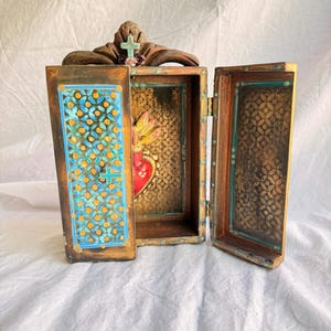 May include: A wooden, open box with a hinged door, featuring a cross and floral accents on top. The interior reveals a heart-shaped design with flames. The box has gold and turquoise details.