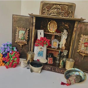 May include: A wooden cabinet with open doors, displaying religious items. Inside are small framed pictures, a statue, and a small book. Outside are colorful bouquets of flowers, a bowl, and a decorative shell. The cabinet has a carved floral design.