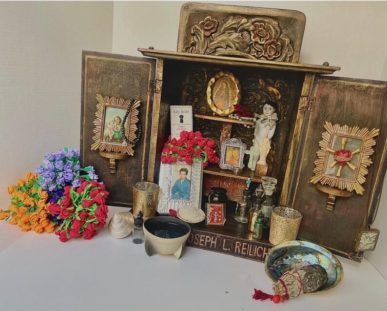 May include: A wooden cabinet with open doors, displaying religious items. Inside are small framed pictures, a statue, and a small book. Outside are colorful bouquets of flowers, a bowl, and a decorative shell. The cabinet has a carved floral design.