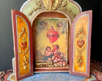 May include: A small wooden shrine with two open doors, featuring a painted image of the Sacred Heart of Jesus with two angels. The shrine is decorated with gold leaf and has a blue and gold patterned base.