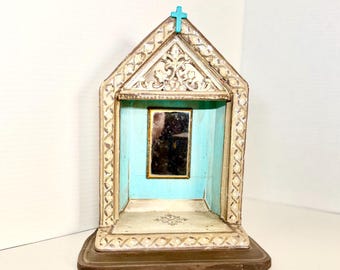 May include: A decorative wooden shadow box with a turquoise interior and a small mirror. The exterior is painted white with ornate carvings and a small blue cross at the top. The base is brown.