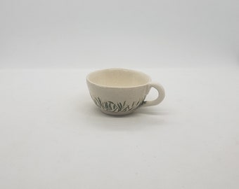 May include: A small, off-white ceramic teacup with a handle. The cup features a hand-painted design of green grass around the lower portion. The interior of the cup is a lighter shade of off-white.