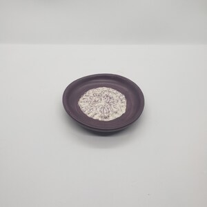May include: A small, round, purple ceramic dish with a textured, off-white floral design in the center. The dish has a slightly raised edge and sits on a white surface. This decorative dish is ideal for holding small items.
