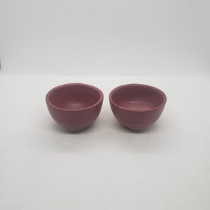 May include: Two small, handmade ceramic bowls. The bowls are a deep reddish-pink color, with a slightly rough texture. The interior of each bowl is a lighter shade of the same color. The bowls are set against a white background.