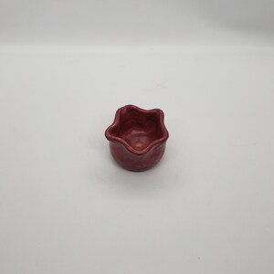 May include: A small, handmade, maroon-colored ceramic bowl with a unique, wavy rim. The bowl has a rounded base and a textured surface, perfect for holding small items. The bowl is likely used for serving dips, sauces, or small snacks.