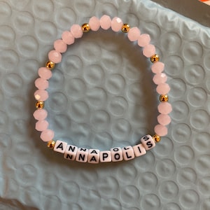 May include: A pink beaded bracelet with gold spacers and white letter beads spelling "ANNAPOLIS".