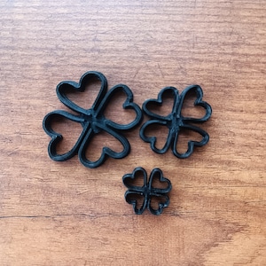 Puede incluir: Tres cortadores de galletas negros en forma de trébol de diferentes tamaños sobre una superficie de madera. Los cortadores son de un material oscuro y tienen un diseño en forma de corazón para cada hoja, ideal para hornear.