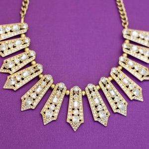 Puede incluir: Un collar de oro con un diseño geométrico adornado con diamantes de imitación transparentes. El collar está sobre un fondo morado.