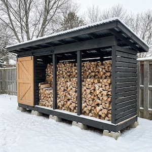 Puede incluir: Un cobertizo de madera negra para leña con techo de metal corrugado y una puerta corredera de madera natural. La estructura está elevada sobre bloques de hormigón y llena de troncos cortados apilados, situada al aire libre en la nieve.