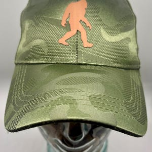 May include: A baseball cap featuring a green camouflage pattern across the crown and brim. Centered on the front panel is a flat, copper-colored silhouette of a Bigfoot figure. The cap has a structured front and stitched detailing on the brim.