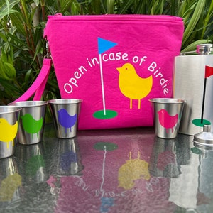 May include: A pink zippered pouch with the text "Open in case of Birdie" and a yellow bird with a golf flag. The pouch is next to a silver flask with a golf flag and a funnel. There are four small silver cups with colorful birds on them.