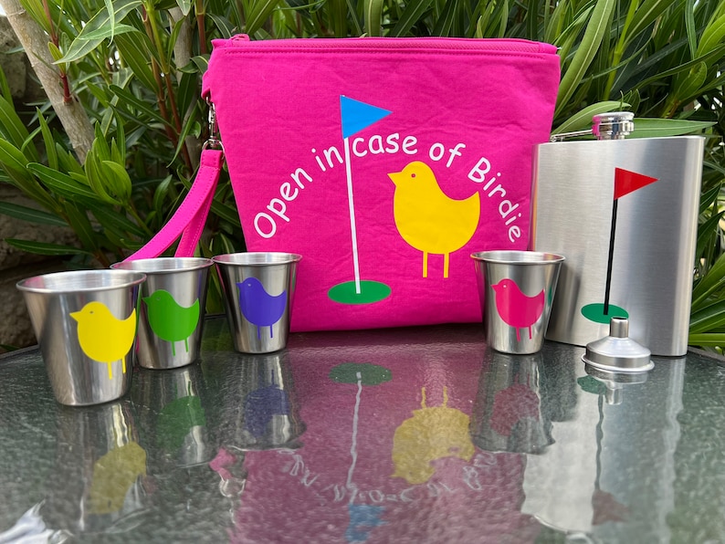 May include: A pink zippered pouch with the text "Open in case of Birdie" and a yellow bird with a golf flag. The pouch is next to a silver flask with a golf flag and a funnel. There are four small silver cups with colorful birds on them.