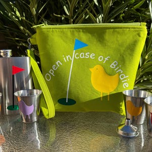 May include: A green zippered pouch with the text "Open in case of Birdie" and a yellow bird illustration. The pouch is next to a silver flask and four silver shot glasses with colorful bird illustrations.