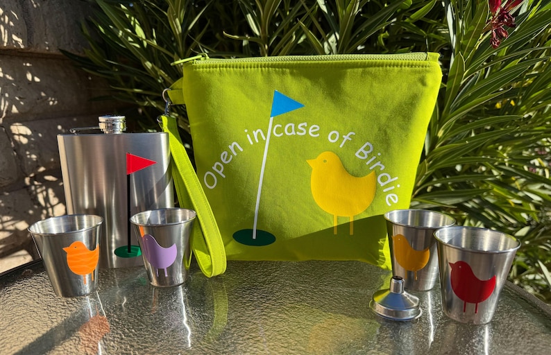 May include: A green zippered pouch with the text "Open in case of Birdie" and a yellow bird illustration. The pouch is next to a silver flask and four silver shot glasses with colorful bird illustrations.