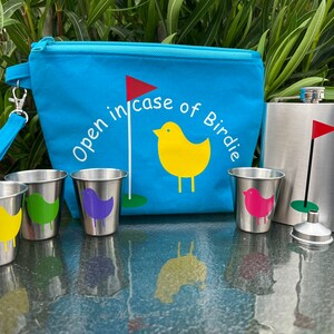 May include: A blue zippered pouch with the text "Open in case of Birdie" and a yellow bird with a red flag on a golf course. The pouch is next to a silver flask with a red flag on a golf course. There are four small silver cups with different colored birds on them.
