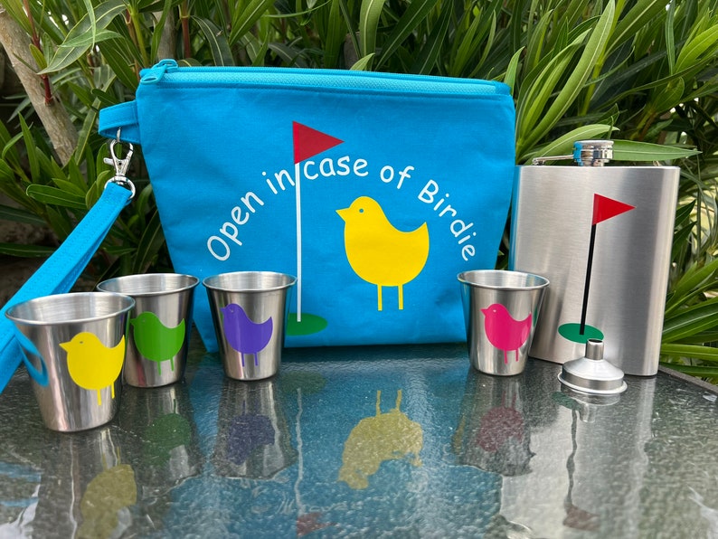 May include: A blue zippered pouch with the text "Open in case of Birdie" and a yellow bird with a red flag on a golf course. The pouch is next to a silver flask with a red flag on a golf course. There are four small silver cups with different colored birds on them.