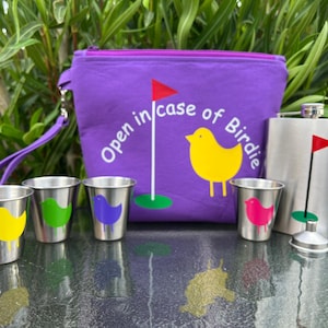 May include: A purple fabric pouch with a zipper closure and a white embroidered design that says "Open in case of Birdie". The pouch has a strap and is next to a silver flask with a golf flag design. There are four small silver cups with colorful bird designs.