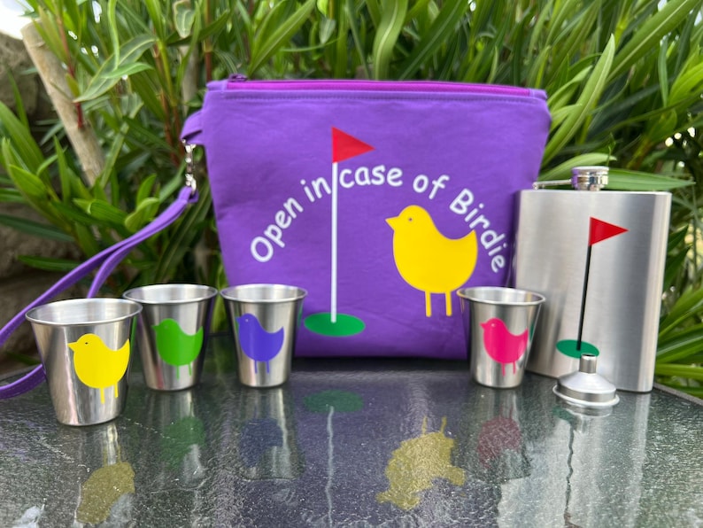 May include: A purple fabric pouch with a zipper closure and a white embroidered design that says "Open in case of Birdie". The pouch has a strap and is next to a silver flask with a golf flag design. There are four small silver cups with colorful bird designs.
