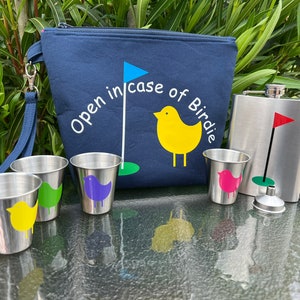 May include: A blue canvas bag with a zipper closure and a handle. The bag has the text "Open in case of Birdie" embroidered on it. The bag is next to a silver flask with a green golf flag and a green and white golf ball. There are four small silver cups with colorful bird designs on them.