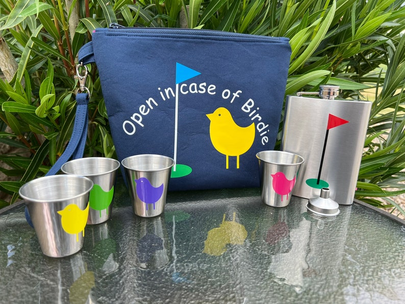 May include: A blue canvas bag with a zipper closure and a handle. The bag has the text "Open in case of Birdie" embroidered on it. The bag is next to a silver flask with a green golf flag and a green and white golf ball. There are four small silver cups with colorful bird designs on them.