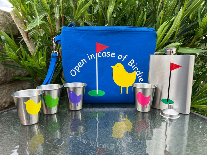 May include: A blue golf-themed pouch with the text "Open in case of Birdie" and a yellow bird graphic. The pouch is next to a silver flask with a red golf flag graphic. There are four small silver cups with colorful bird graphics.
