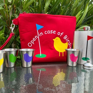 May include: A red golf-themed pouch with the text "Open in case of Birdie" and a blue flag, along with four small stainless steel cups with colorful bird designs and a stainless steel flask with a golf flag design.