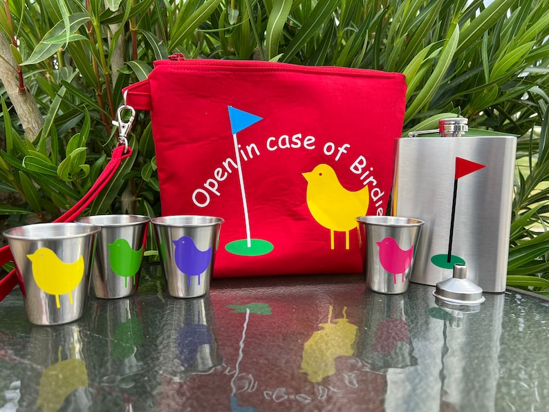 May include: A red golf-themed pouch with the text "Open in case of Birdie" and a blue flag, along with four small stainless steel cups with colorful bird designs and a stainless steel flask with a golf flag design.