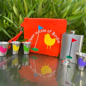 May include: A red pouch with the text "Open in case of Birdie" and a yellow bird graphic. The pouch is next to a silver flask with a golf flag graphic. There are four small silver shot glasses with colorful bird graphics.