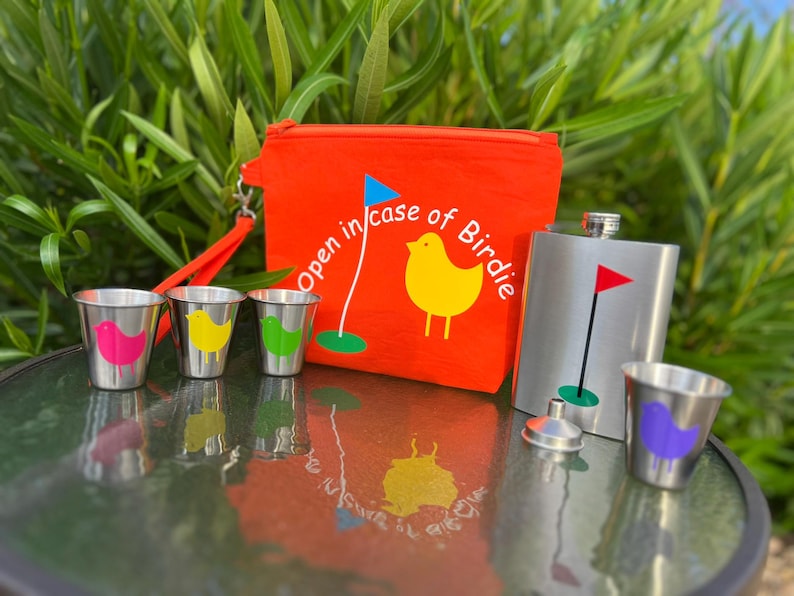 May include: A red pouch with the text "Open in case of Birdie" and a yellow bird graphic. The pouch is next to a silver flask with a golf flag graphic. There are four small silver shot glasses with colorful bird graphics.
